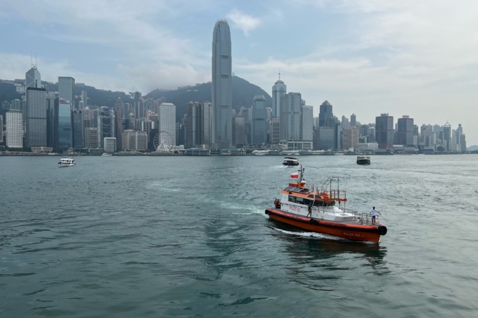 A red and white rescue boat navigates the waters of Victoria Harbour with the Hong Kong skyline, including the International Finance Centre, towering in the background under a partly cloudy sky.